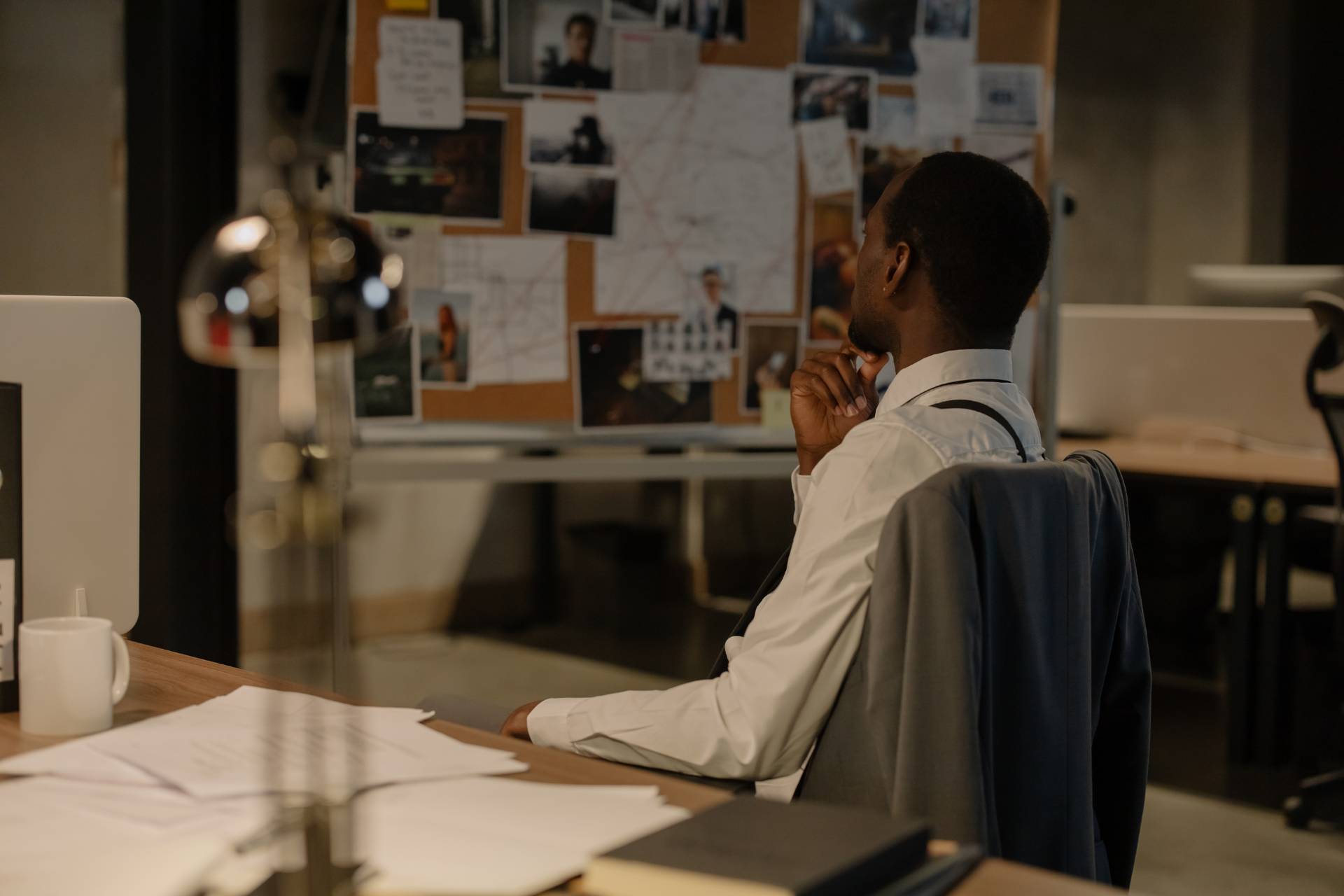 A man in an office sits at his desk, looking thoughtfully at a bulletin board covered with photos, documents, and a large map—perhaps analyzing data for background checks or screening services.
