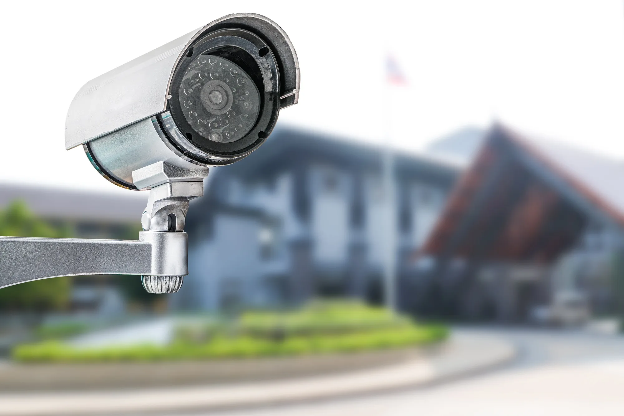 A security camera mounted on a metal bracket with a blurred building and flag in the background, indicative of modern AI surveillance services in Georgia.