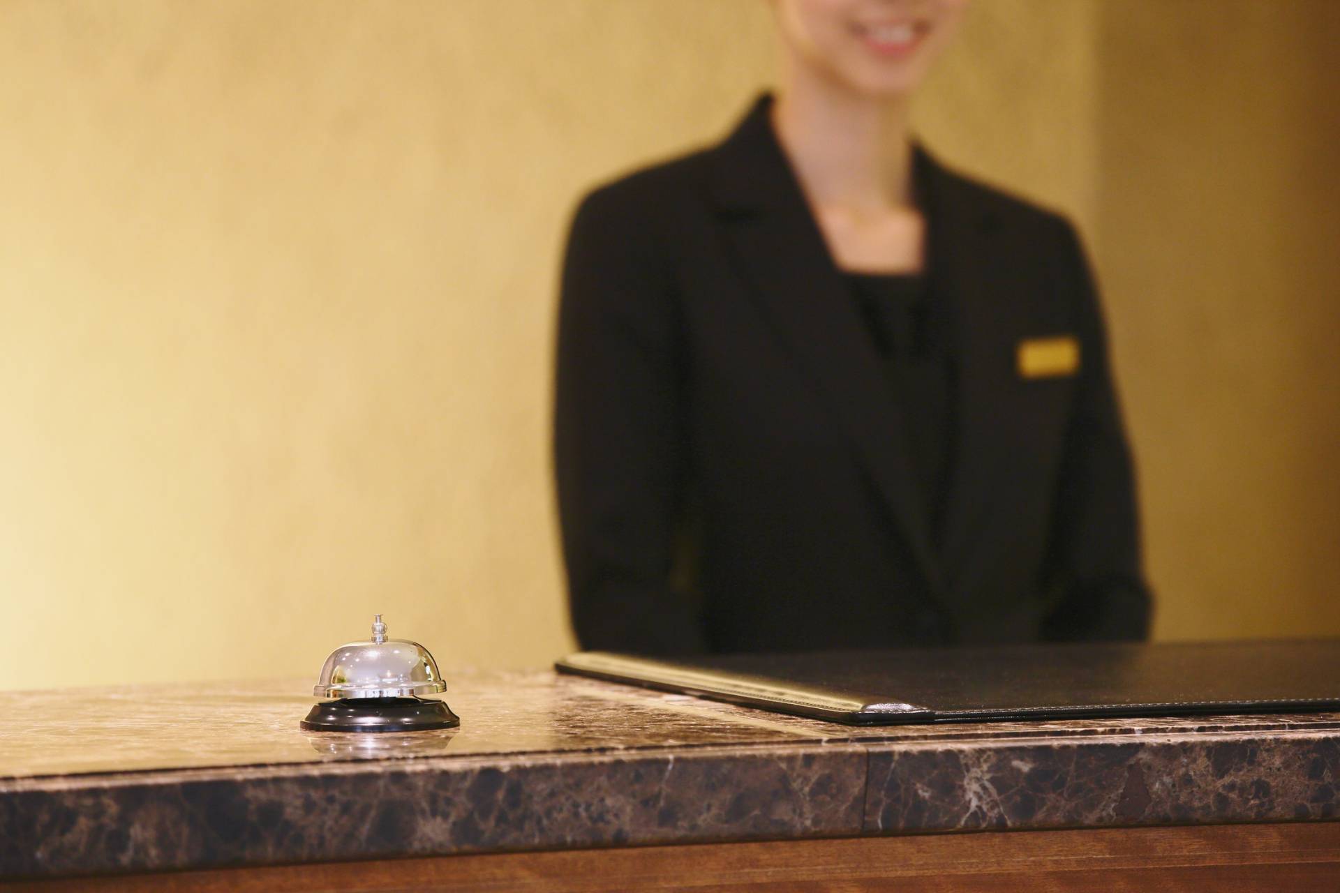 A hotel receptionist in a black uniform stands behind a marble counter with a service bell and black folder, ready to offer concierge services along with top-tier security.