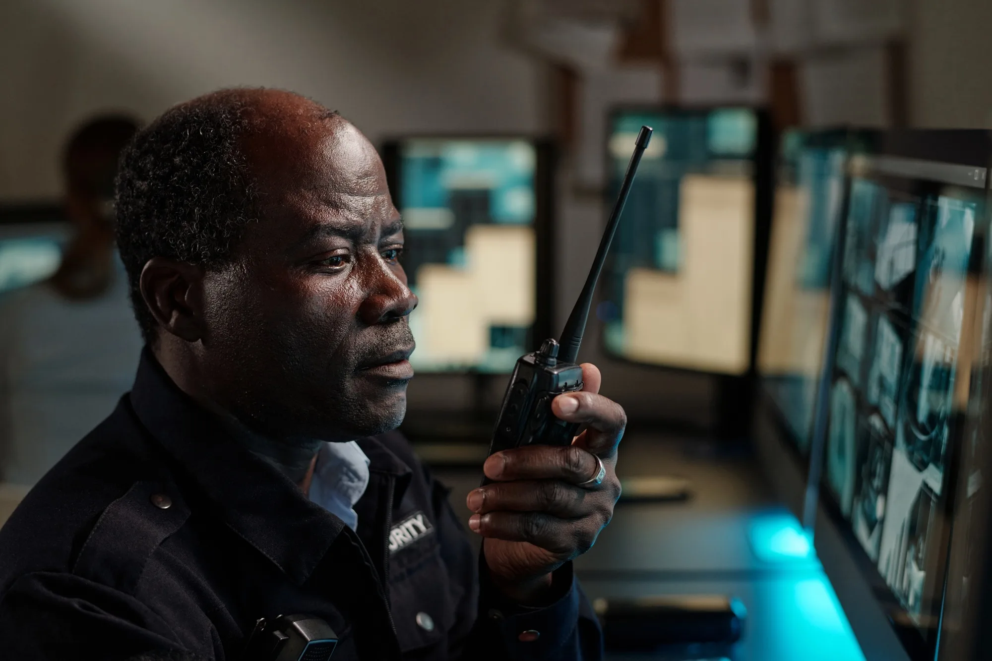 A security guard holds a walkie-talkie and monitors multiple surveillance screens in a control room, representative of the vigilance found in off-duty police security in Georgia.