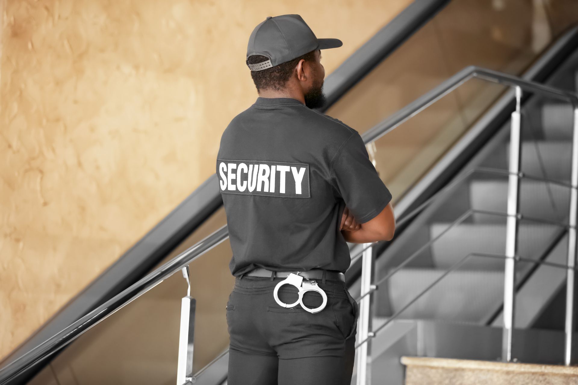 A security guard in uniform stands with arms crossed near a staircase, with handcuffs attached to their belt.