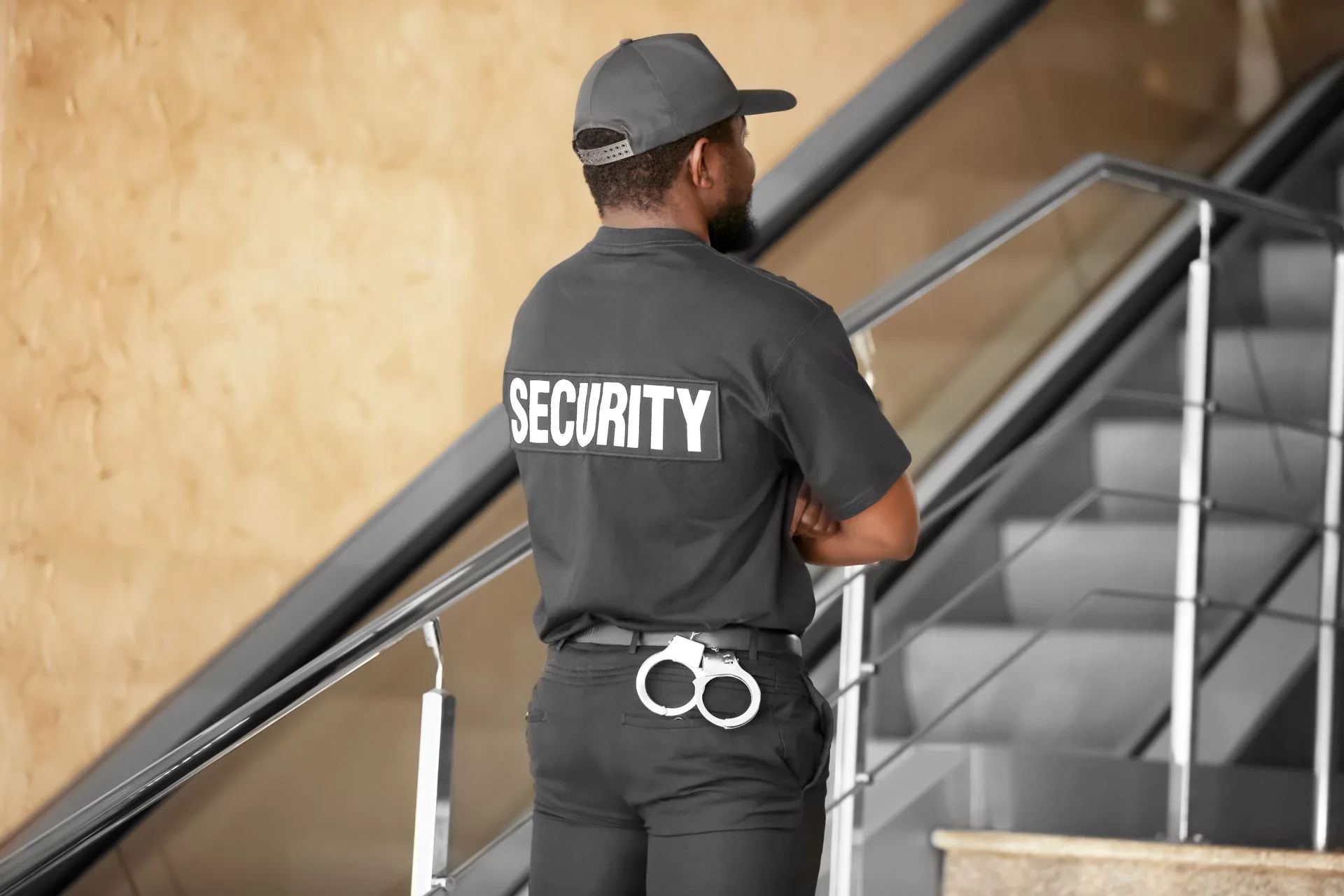 A security guard in uniform stands with arms crossed near a staircase, with handcuffs attached to their belt.
