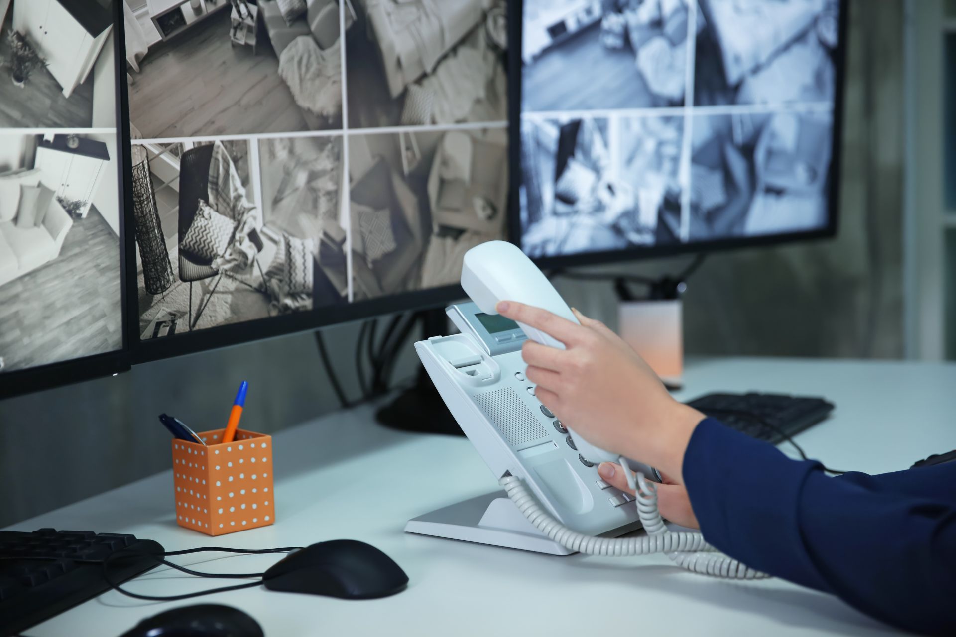 Person holding a landline phone at a desk with computer monitors displaying multiple security camera feeds in the background.