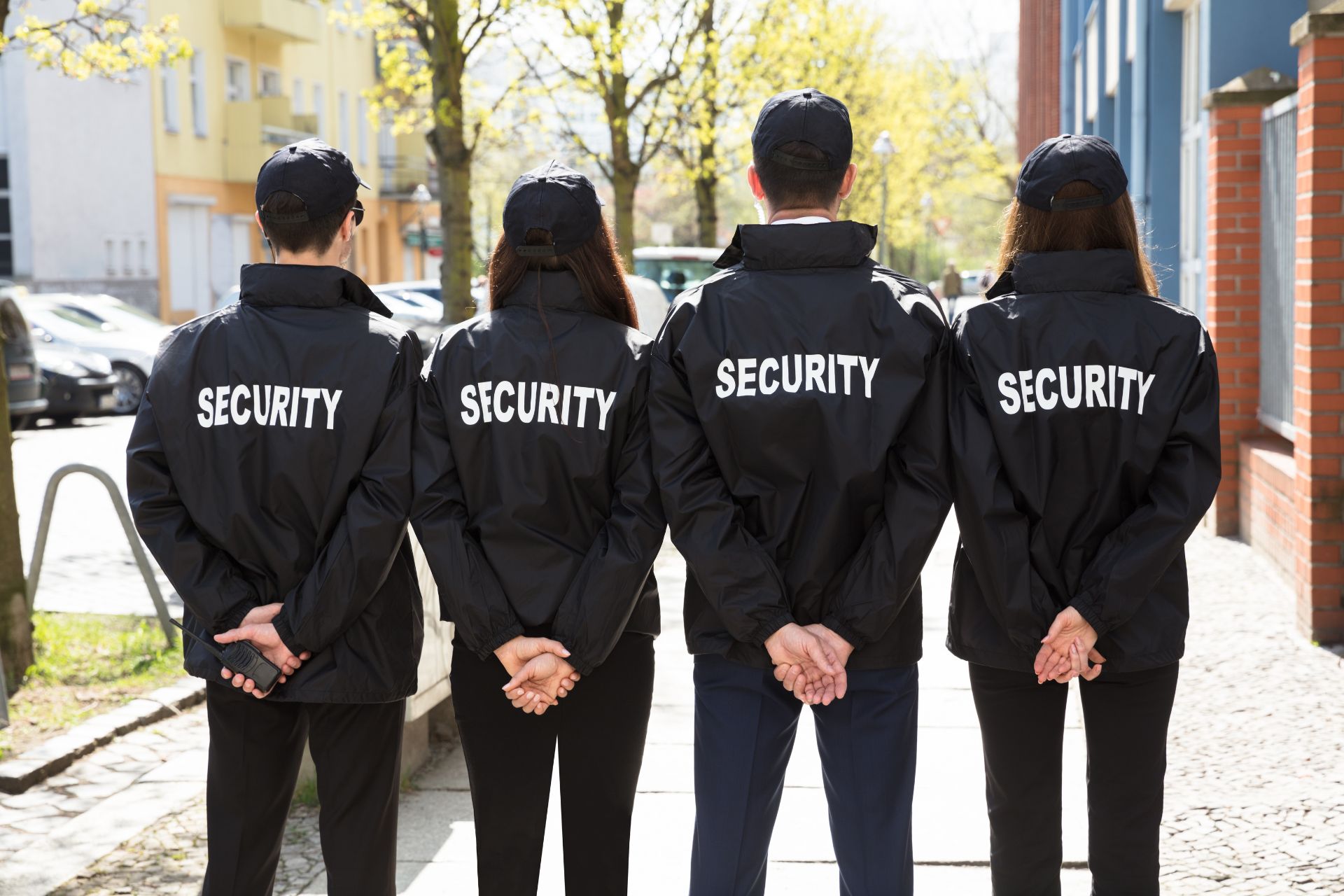 Four security guards in uniform stand outdoors with their backs to the camera, hands behind their backs, on a sidewalk lined with buildings and trees.