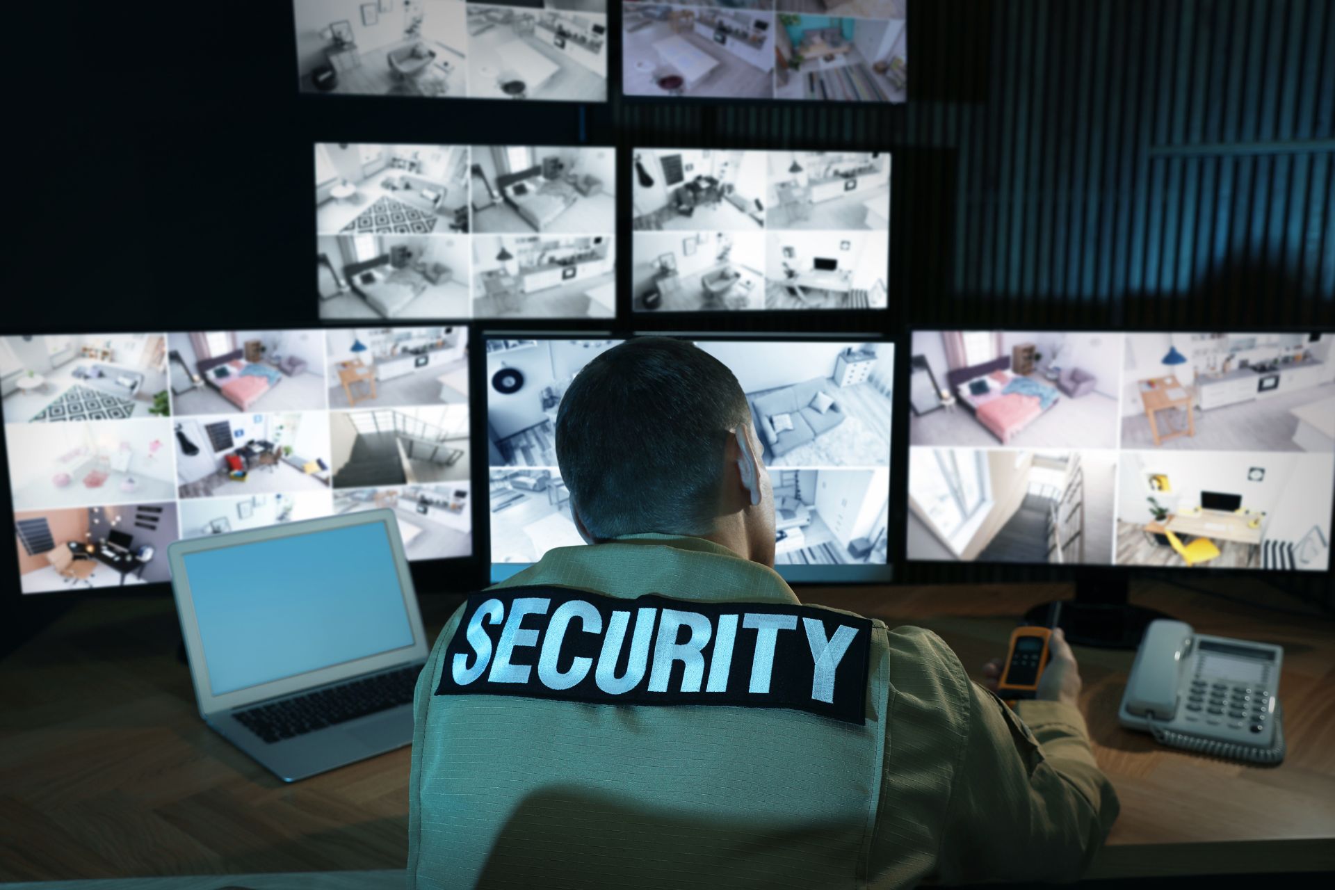 A security guard monitors multiple surveillance camera feeds on screens in a control room, with a laptop and phone on the desk.