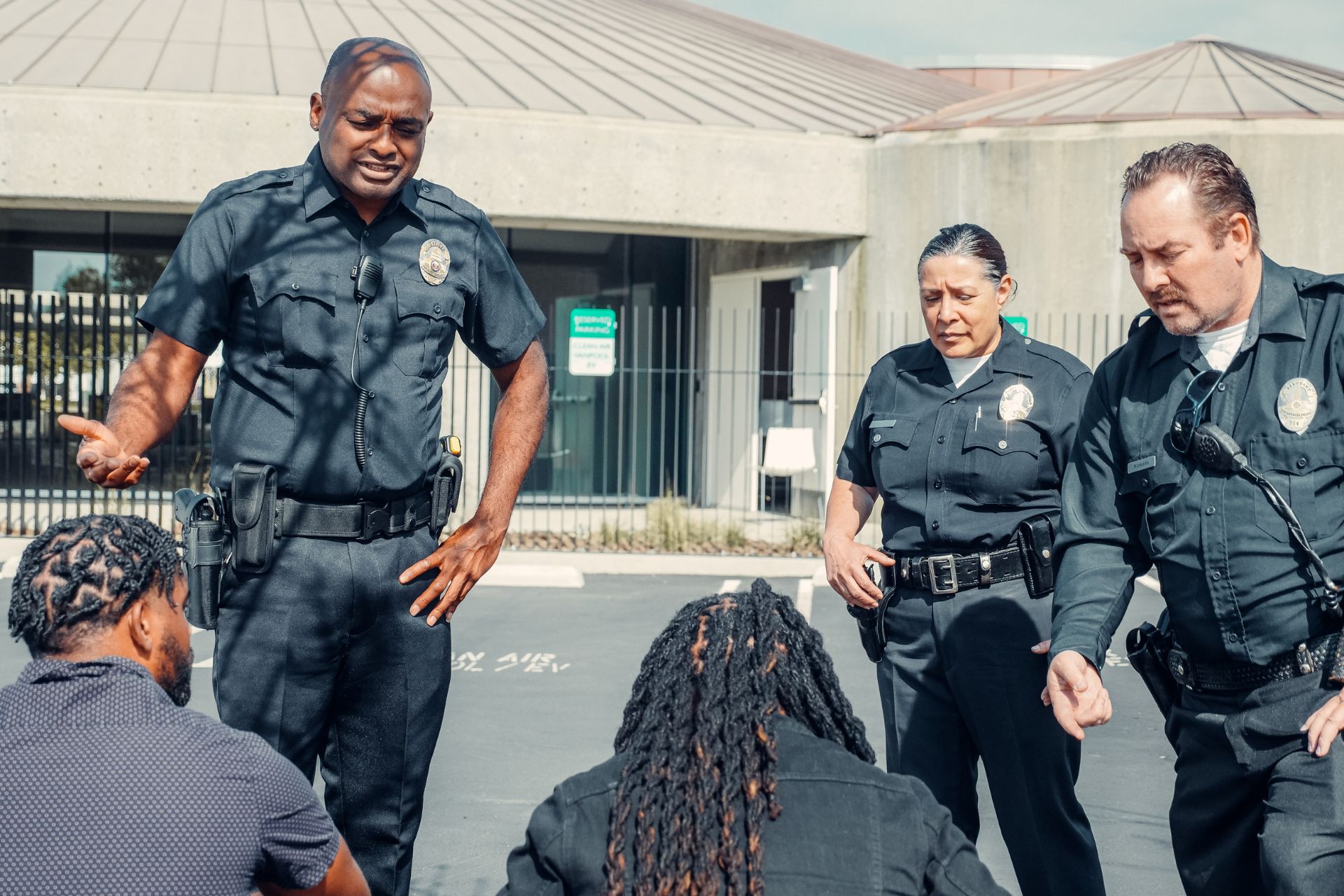 Four police officers stand and talk to two seated individuals with their backs to the camera in a parking lot outside a building.
