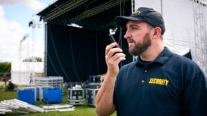 Security Team At Outdoor Stage For Music Festival Or Concert Talking Into Radio