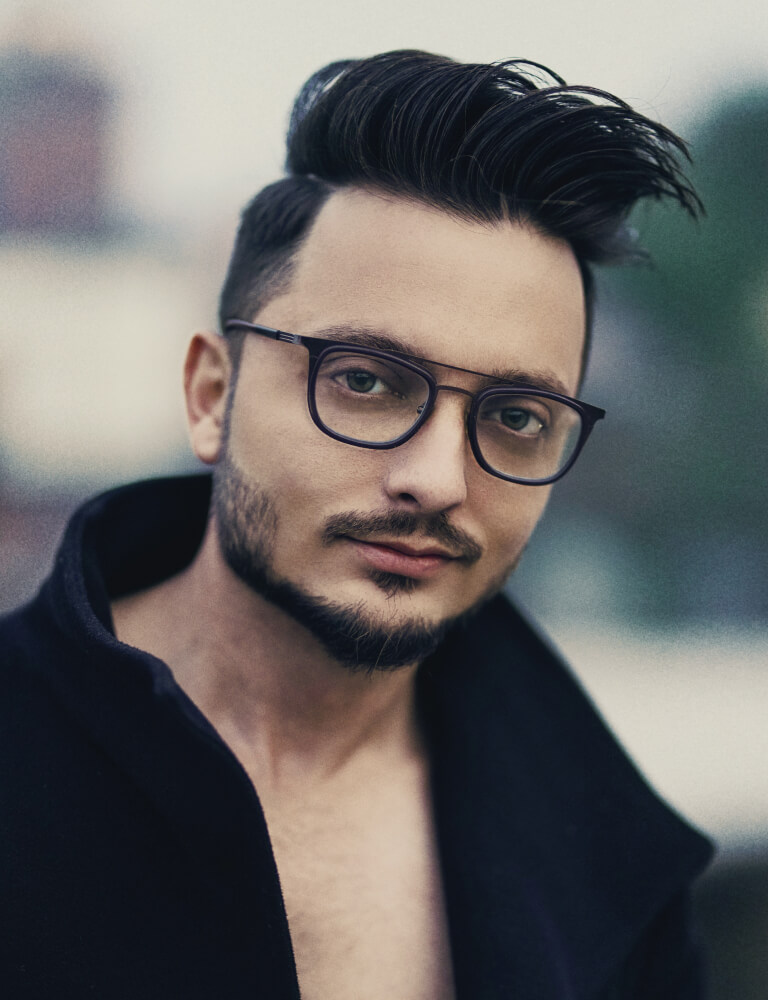 A man with styled dark hair, trimmed beard, and glasses wearing a dark coat looks directly at the camera.