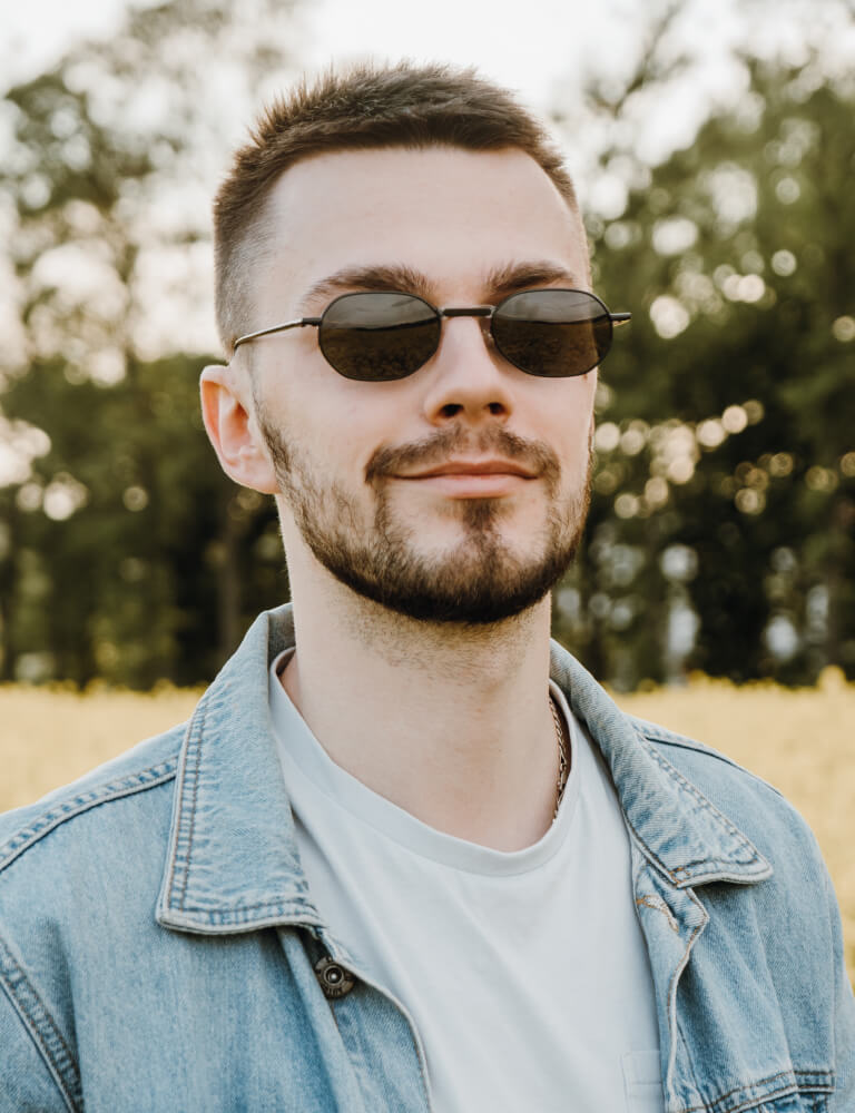 A man with short hair, a beard, and sunglasses wearing a light denim jacket and white shirt stands outdoors with blurred trees and grass in the background.