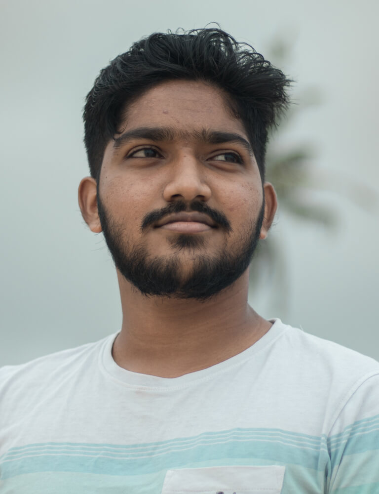 A young man with a beard and short hair looks to the side while standing outdoors, wearing a white t-shirt. A blurred palm tree is visible in the background.