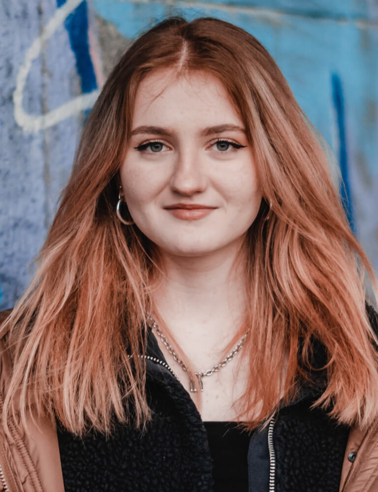 A young woman with long reddish-blonde hair, wearing a brown jacket and layered necklaces, stands in front of a blue graffiti wall.