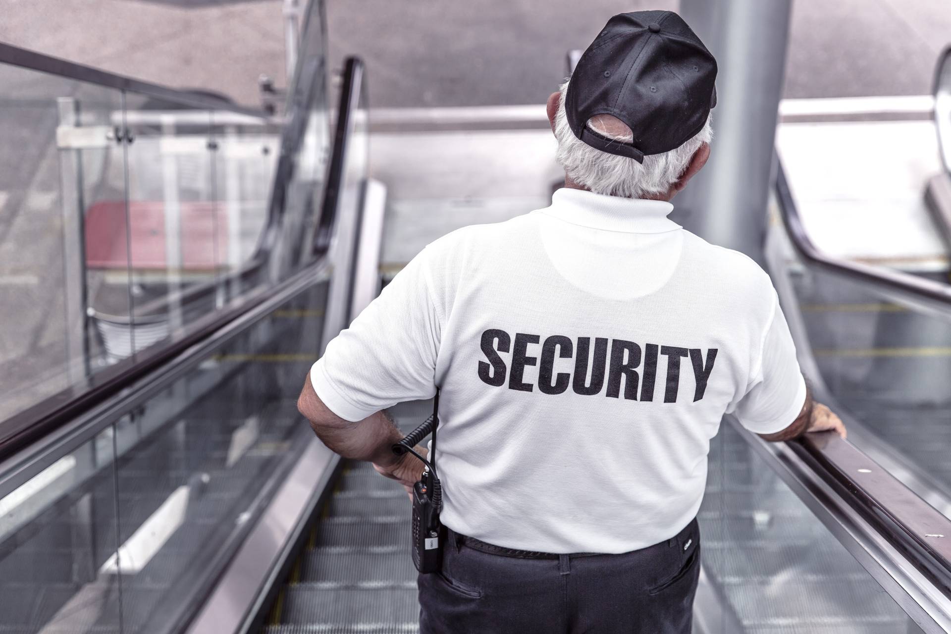 An unarmed security guard in a white shirt and black cap stands on an escalator, facing away, with "SECURITY" written on the back of the shirt.