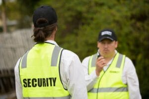 Two unarmed security guards in yellow vests, one facing away and the other speaking into a radio, stand outdoors near a fence and trees.