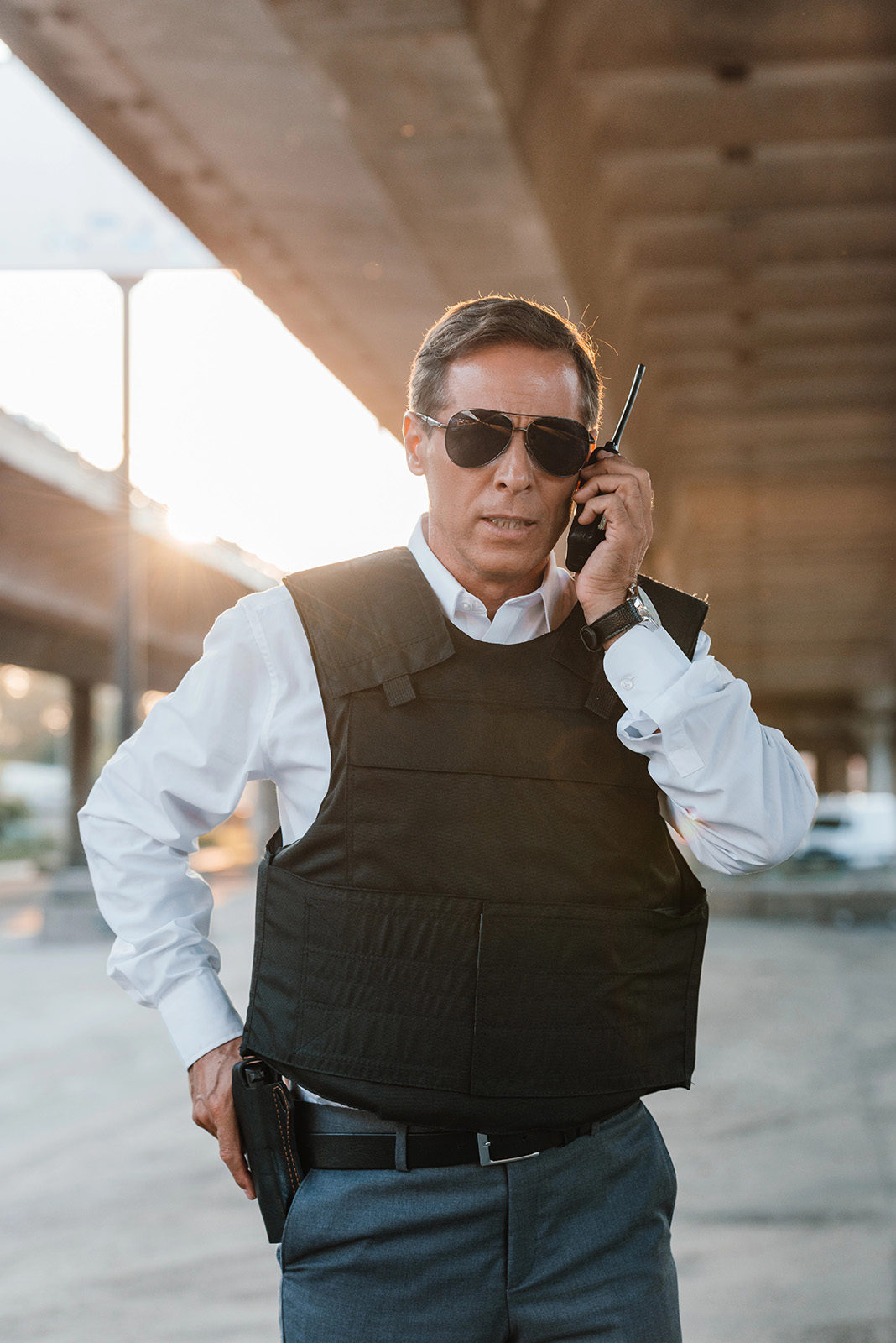 Man in a white shirt, black vest, and sunglasses—an off-duty police security in Georgia—talks on a walkie-talkie under an overpass.