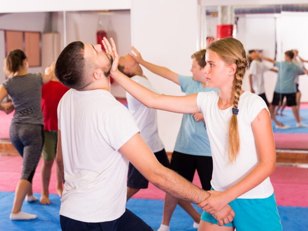 A group of people practice Self Defense Basics in a gym; a girl in the foreground uses her hand to push away a partner&rsquo;s face.