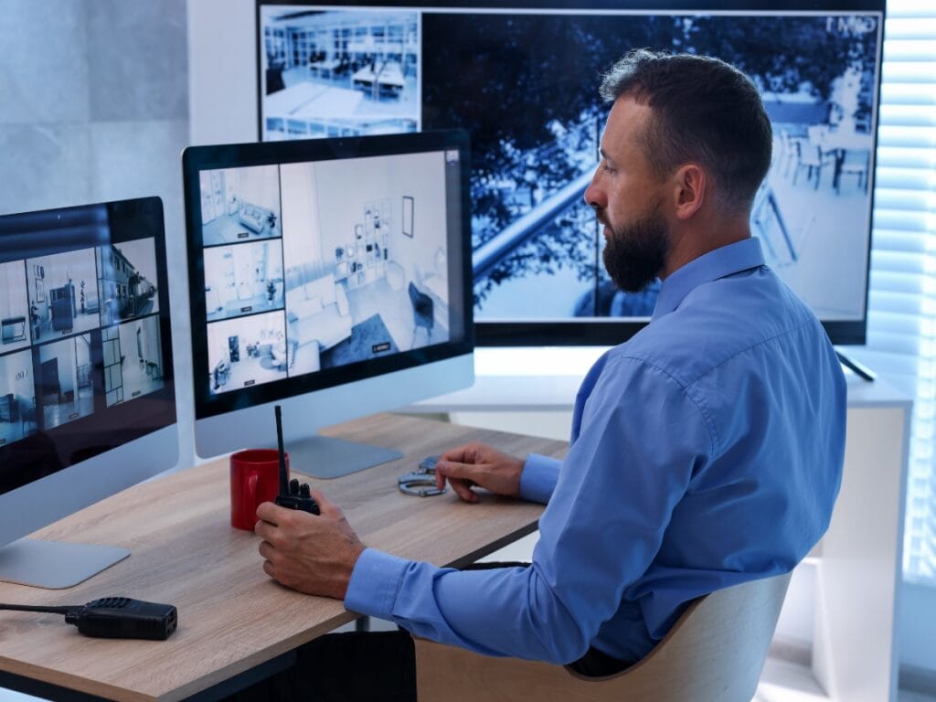 A man in a blue shirt monitors multiple Smart Home Security camera feeds on computer screens while holding a walkie-talkie at his desk.