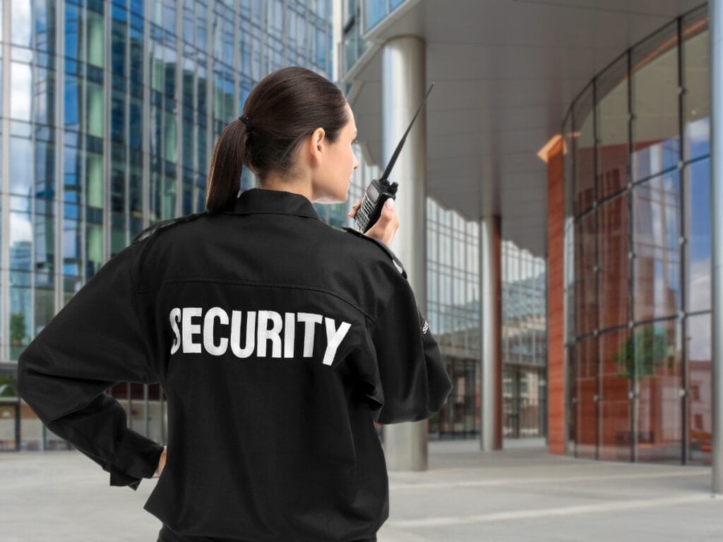 A security officer in uniform stands outside a modern glass building, holding a two-way radio and monitoring the area during private parties.