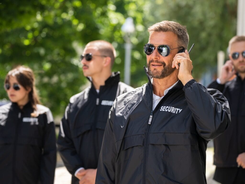 Four security personnel wearing sunglasses and black jackets stand outdoors, with one man in front speaking into an earpiece&mdash;professional protection often seen at exclusive private parties. Greenery is visible in the background.