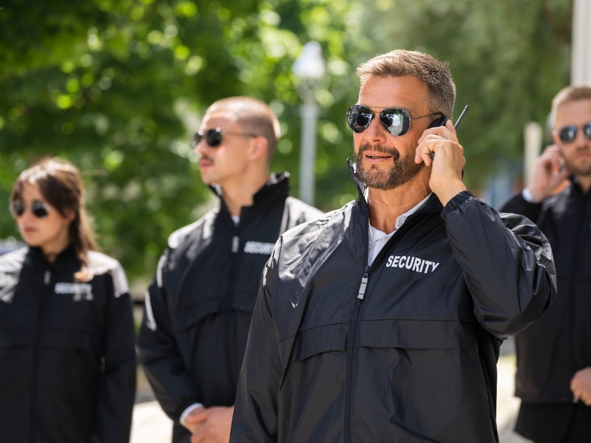 Four security personnel wearing sunglasses and black jackets stand outdoors, with one man in front speaking into an earpiece—professional protection often seen at exclusive private parties. Greenery is visible in the background.
