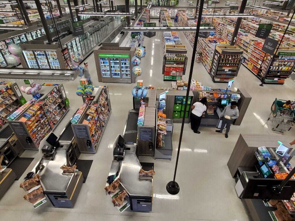 Aerial view of a grocery store with self-checkout stations equipped for retail loss prevention, two people using a kiosk, and aisles stocked with various products in the background.