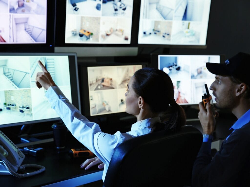 Two people in a security control room monitor multiple surveillance camera feeds for retail loss prevention; one points at a screen while the other holds a walkie-talkie.