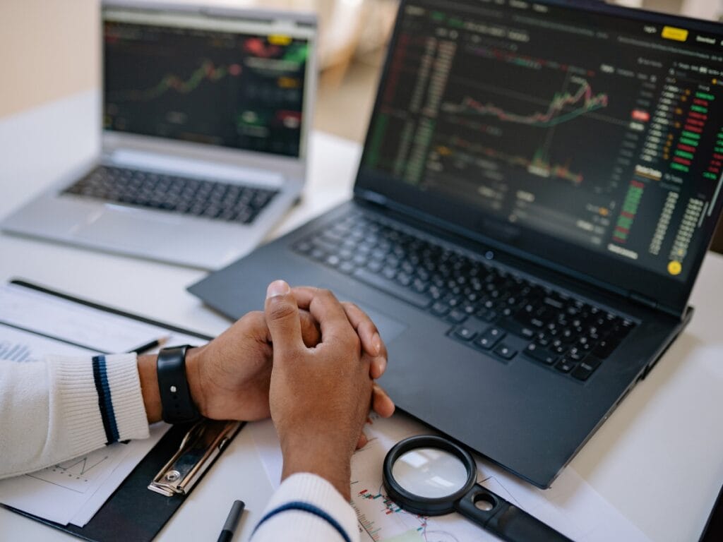 Person sits at a desk with folded hands in front of two laptops displaying stock market charts and data, alongside papers, a magnifying glass, a pen, and notes on Digital Forensics Basics for investigating financial activity.