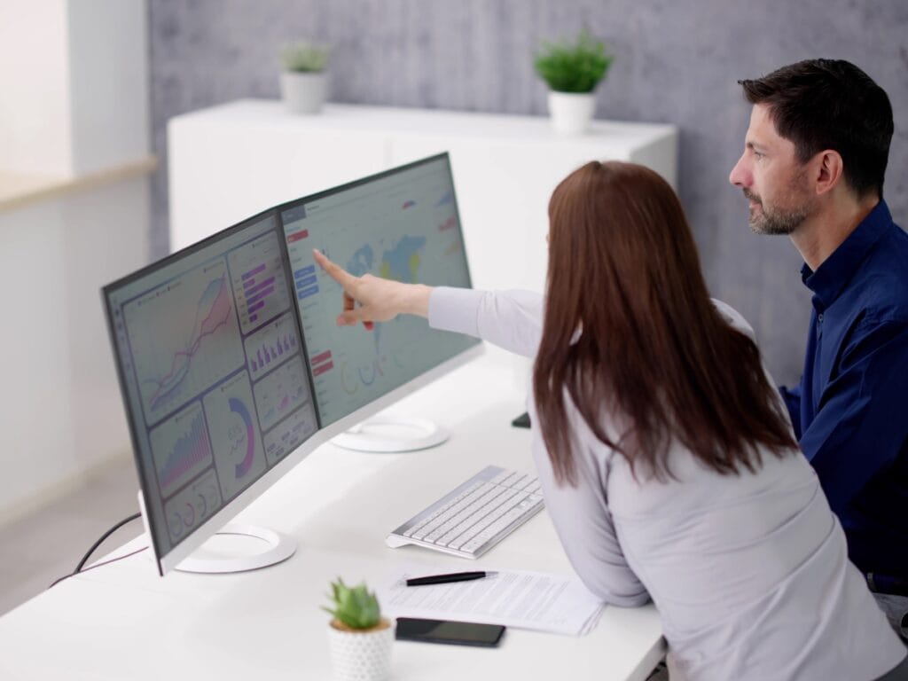 Two people sit at a desk looking at data and charts on dual computer monitors, discussing Digital Forensics Basics as one person points at the screen.