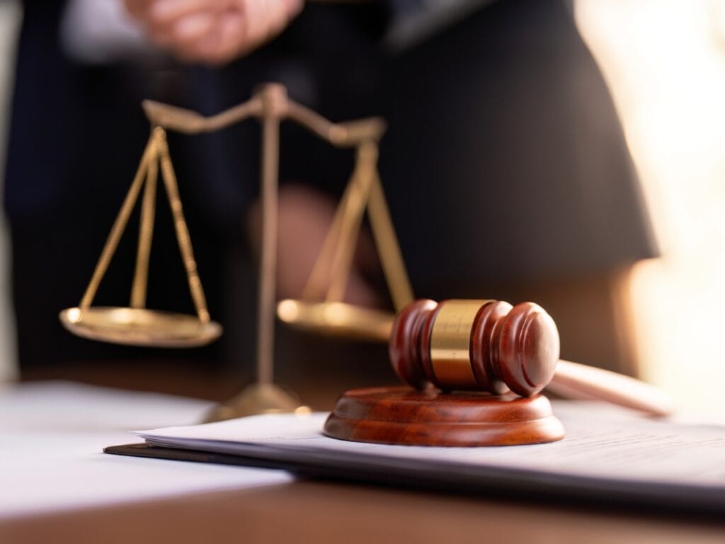 A wooden gavel rests on a sound block in front of a stack of papers labeled “Digital Forensics Basics,” with brass balance scales and a blurred figure in a suit in the background.