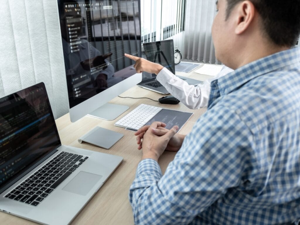 Two people at a desk with multiple computers and laptops, discussing code and Digital Forensics Basics displayed on large monitor screens in an office setting.