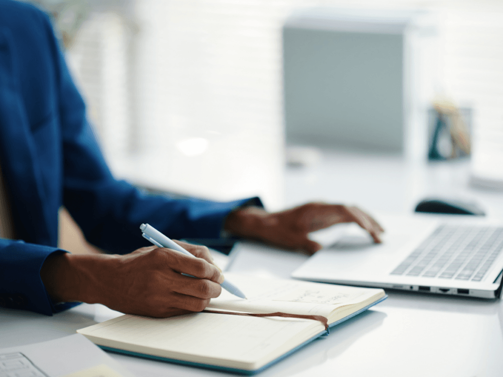 Person in a suit engaged in report writing, jotting notes in a notebook with a pen while using a laptop at a desk in a bright office environment.