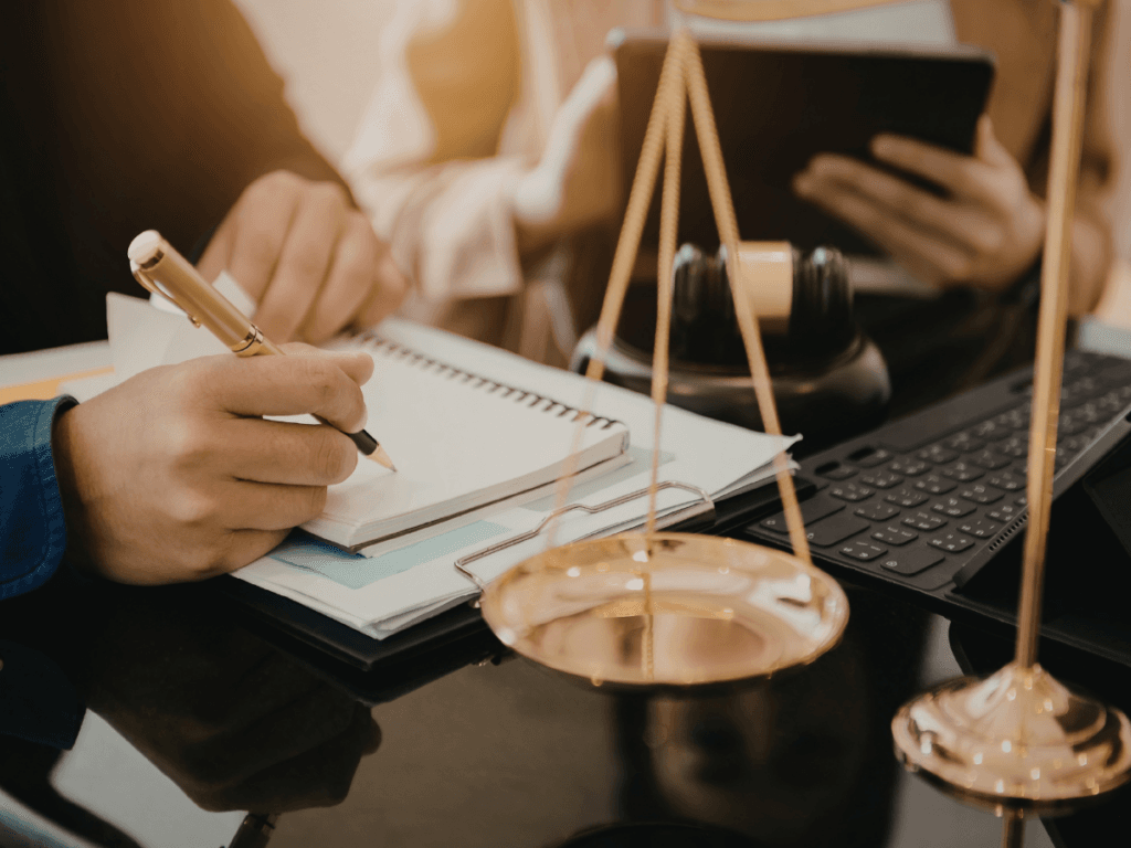 Two people at a desk with legal documents, a gavel, scales of justice, a notebook, tablet, and keyboard visible. One person is writing notes for report writing.