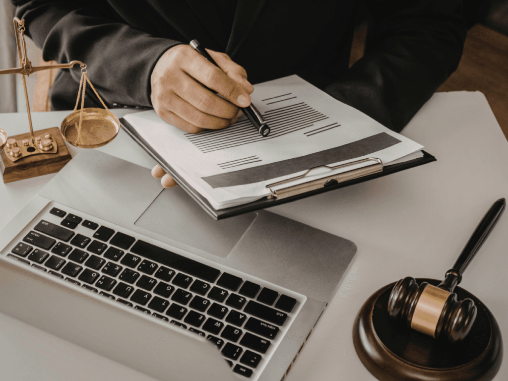 A person in a suit reviews and marks documents on a clipboard at a desk with a laptop, gavel, scales of justice, and focuses on report writing.
