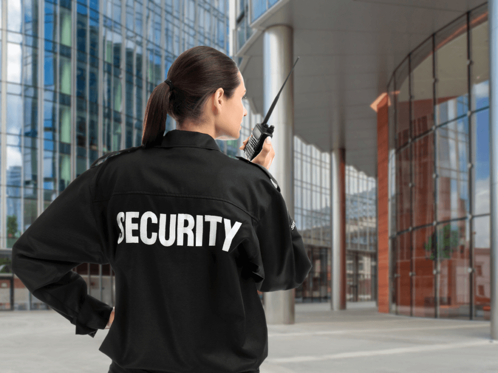 A security guard in uniform stands outside a modern building, holding a walkie-talkie and preparing for report writing while watching the glass entrance.