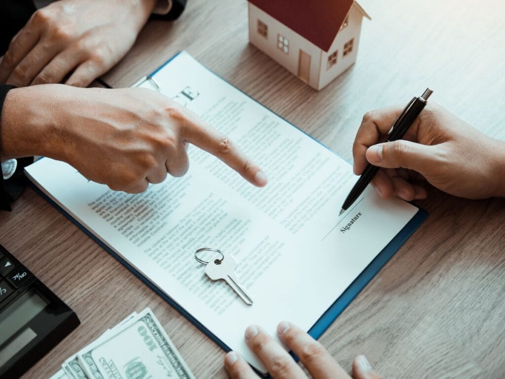 A person signs a document on a clipboard while another points at it; a house model, keys, and dollar bills are on the table, highlighting the importance of background screening in real estate transactions.