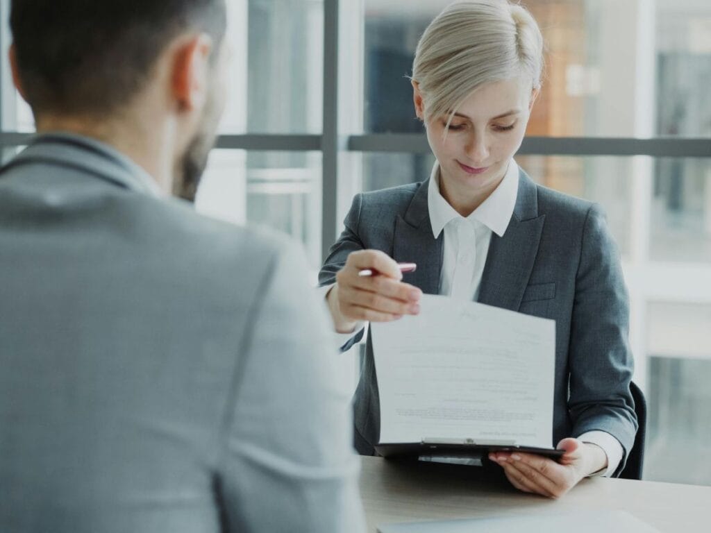A woman in a business suit hands a document to another person across a table in a modern office setting, highlighting the importance of background screening during professional meetings.