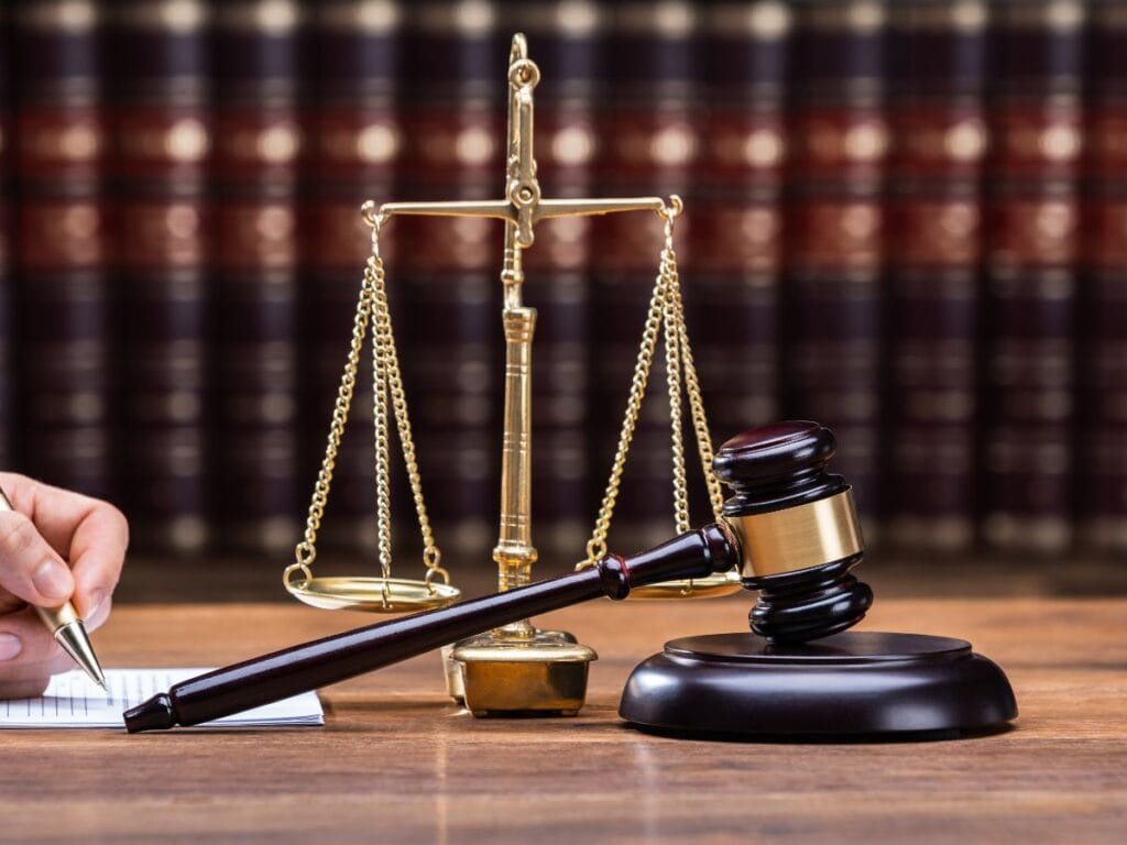 A gavel and scales of justice on a wooden desk with a hand writing in a notebook; law books and background screening documents are visible in the background.