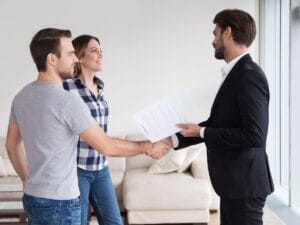 A man in a suit, after completing background screening, hands documents to a casually dressed couple; he shakes hands with the man while the woman stands beside them, smiling, in a living room.