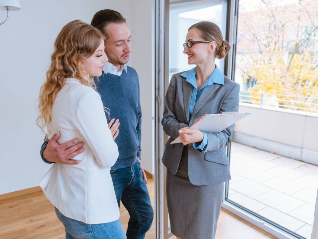 A real estate agent, after background screening, shows a young couple an apartment, standing together indoors near a large window with a clipboard in hand.