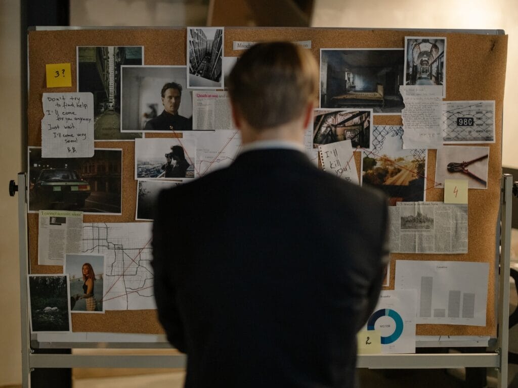A person in a suit stands facing a corkboard covered with photographs, notes, maps, and newspaper clippings—string connecting various points as they investigate a tangled web of possible infidelity.