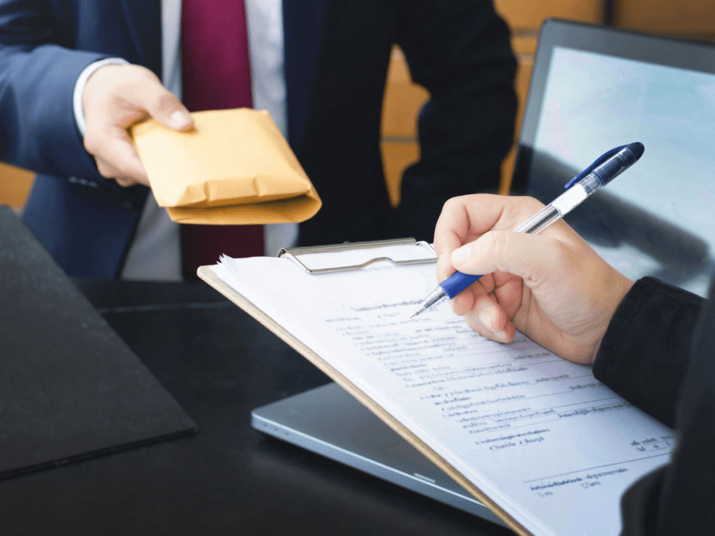 Two people in business attire exchange a brown envelope while one writes on a clipboard with documents, next to a laptop on a desk—a scene often scrutinized during corporate fraud detection investigations.