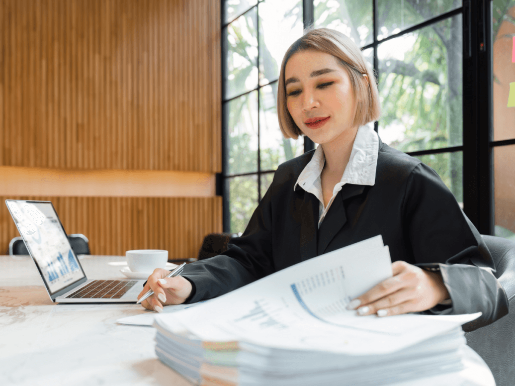 A woman in business attire reviews documents at a desk with a laptop and coffee cup, focused on corporate fraud detection, with large windows illuminating the background.