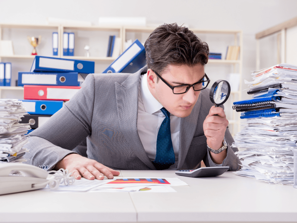A man in a suit uses a magnifying glass to examine a calculator at a cluttered desk with large stacks of paperwork and binders, illustrating the meticulous process of corporate fraud detection.