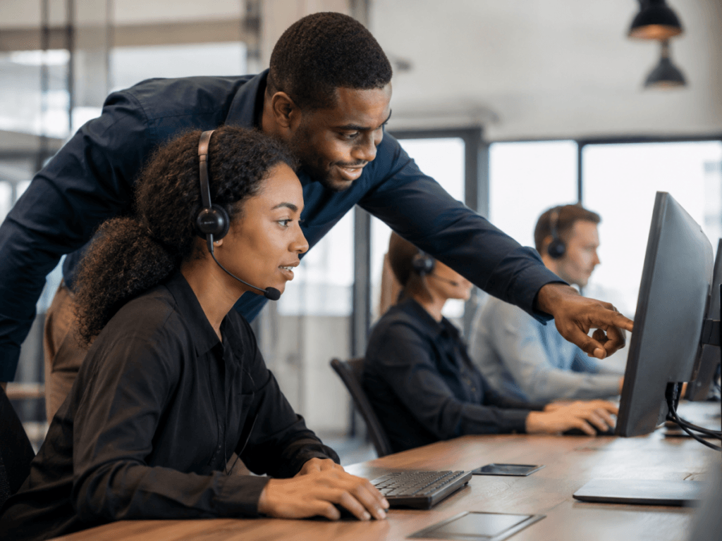 Two Security Customer Service representatives wearing headsets work at computers. One stands and points at the monitor, assisting the other, who is seated. Other colleagues are visible in the background.