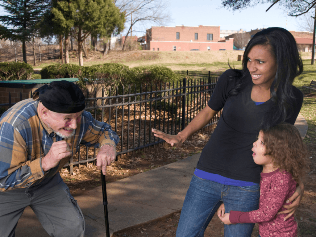 An older man with a cane approaches a woman and a young girl in a park. The woman holds the girl close and extends her hand toward the man, showing caution as if aware of stranger danger.