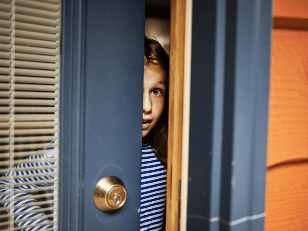A person in a blue and white striped shirt peeks out from behind a partially opened door, looking outside with a hint of caution, aware of stranger danger.