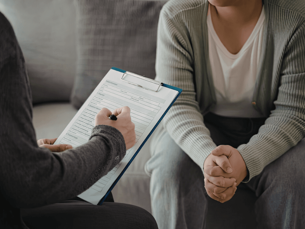 A person fills out a form on a clipboard while sitting across from another, both aware of stranger danger, their hands clasped as they sit together on a sofa.