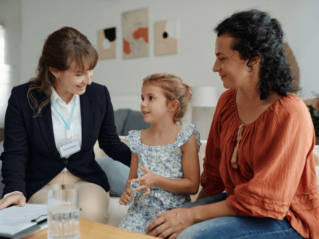 A woman in a suit discusses stranger danger with a young girl and an adult woman, all sitting together indoors and appearing engaged in conversation.