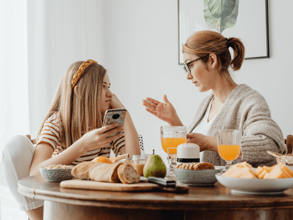 Two people sit at a table with breakfast foods and orange juice; one is holding a phone, while the other gestures, perhaps warning about stranger danger during their morning conversation.