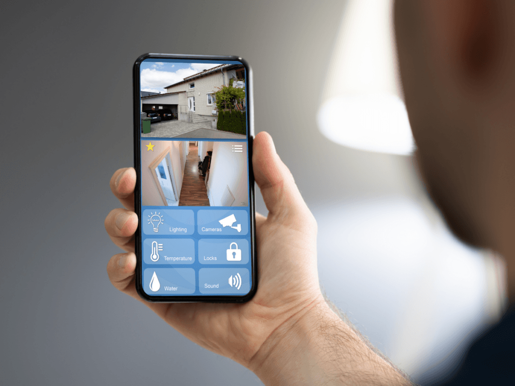 A person holds a smartphone displaying a home security app with live camera feeds and controls, using vacation home security features to monitor lighting, cameras, temperature, locks, water, and sound while away on summer travel.