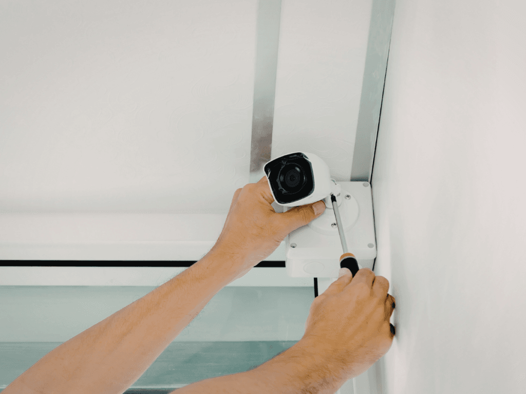 Person installing a security camera on a white ceiling using a screwdriver, following key security system tips to enhance vacation home security before summer travel.
